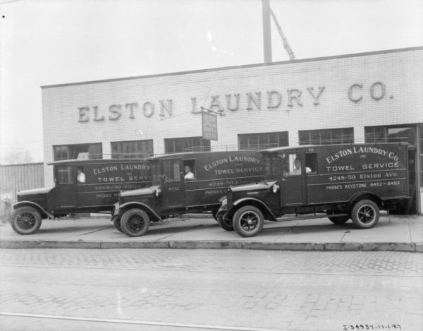 View across street towards three men sitting in the driver's seats of three trucks. They are parked at an angle in front of a commercial building with a sign on the front that reads: "Elson Laundry Co."