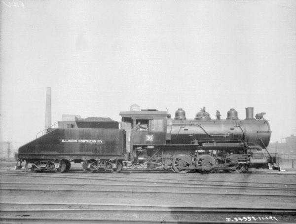 Right side profile view of a locomotive pulling a railroad car with a sign painted on the side that reads: "Illinois Northern Ry." The railroad engineer is sitting in the open window, above the number "16" painted on the side. There are a number of railroad tracks in the foreground. In the background is a smokestack and industrial buildings.