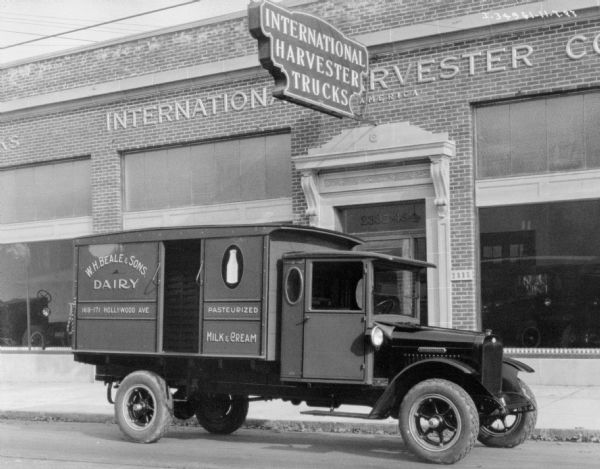 View across street towards a delivery truck parked in front of an International Harvester dealership. The sign painted on the side of the truck reads: "W.H. Beale & Sons Dairy."