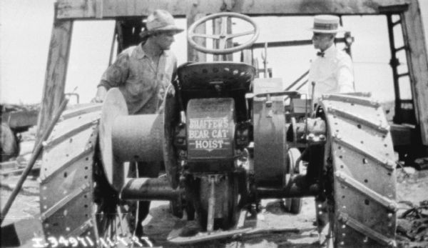 Two men are standing on either side of a Shaffers Bear Cat Hoist and a McCormick tractor. The base of an oil rig is in the background.