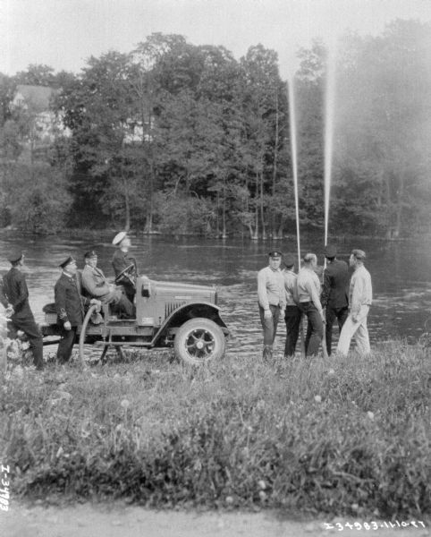 A group of men at a water pump demonstration on the shoreline of a body of water. There is a truck on the left side.