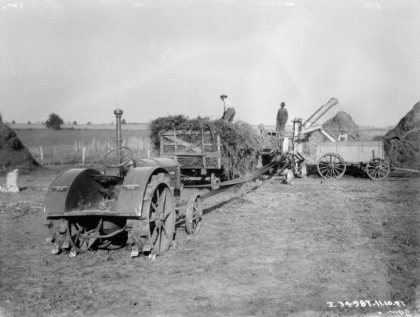 A McCormick-Deering 15-30 tractor is powering a thresher. A man is standing on top of a pile of grain on a wagon, and another man is standing on the thresher. One other man is standing behind a wagon on the right.