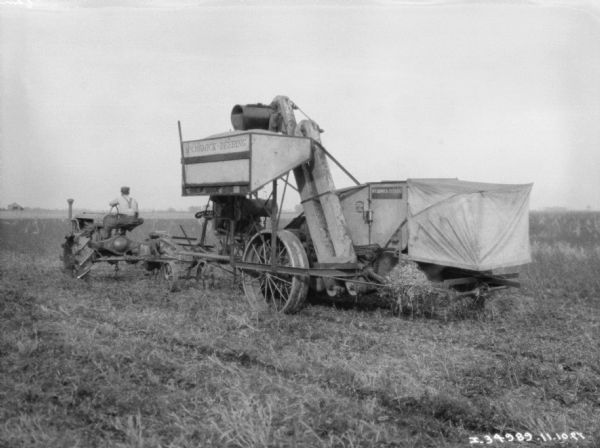 Three-quarter rear view from left of a man driving a tractor-powered harvester thresher in a field.
