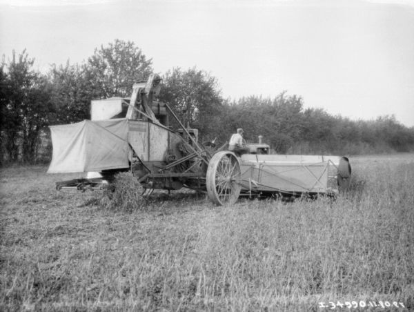 Three-quarter rear view from right of a man driving a tractor pulling a harvester thresher.