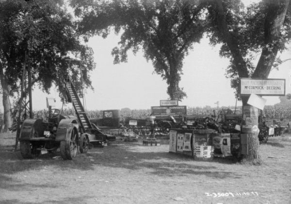 View across lawn towards a display of tractors, trucks, cream separators, and the other implements around a counter with posters and signs. There is a cornfield in the background.