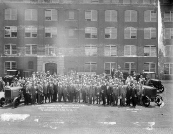 Elevated group portrait of men and woman standing outdoors in front of a factory building. There are trucks and tractors framing the group, who were guests at the International Harvester Company at the McCormick Works in Chicago.