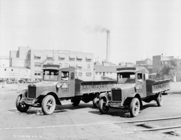 Two men are sitting in the driver's seats of two trucks parked at an angle. The signs painted on the truck read: "T.M. Sinclair & Co., Ltd., Beef and Pork Packers." A building and smokestack are in the far background. Signs on the building read: "Office" and "Sup't. Office."