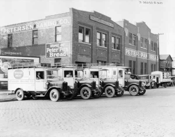 View across cobblestone street towards truck drivers sitting in four delivery trucks in front of a billboard and a brick building. The signs on the building in the background read: "Petersen Baking Co." and "Home of Peter Pan Bread." The billboard has an advertisement for Red Crown Gasoline.