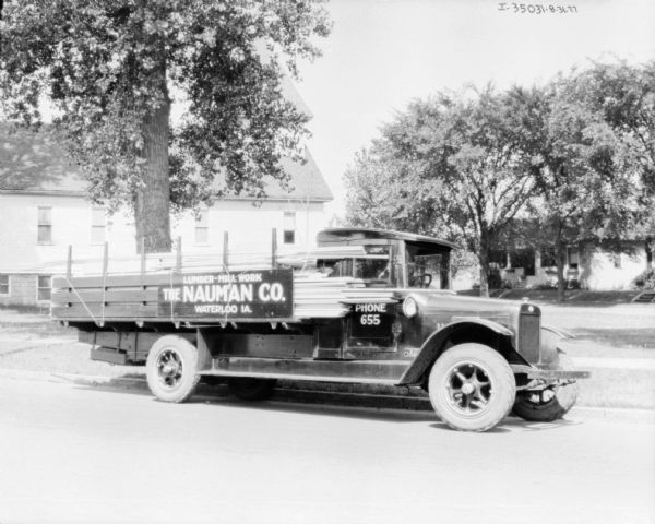 View across street towards a truck parked along the curb. The sign painted on the side of the truck reads: "Lumber-Millwork, The Nauman Co." There is a large building in the background. A stack of lumber extends from the back of the truck bed to the front of the truck, along the side of the passenger side door.
