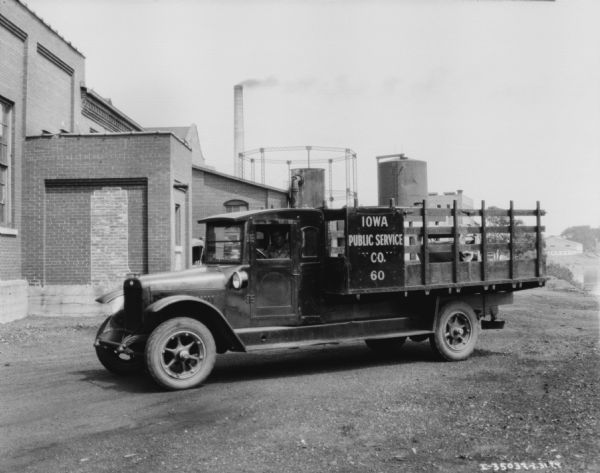 View of a man sitting in the driver's seat of a truck. The sign painted on the truck reads: "Iowa Public Service Co." The truck is parked near a brick industrial building and storage tanks.