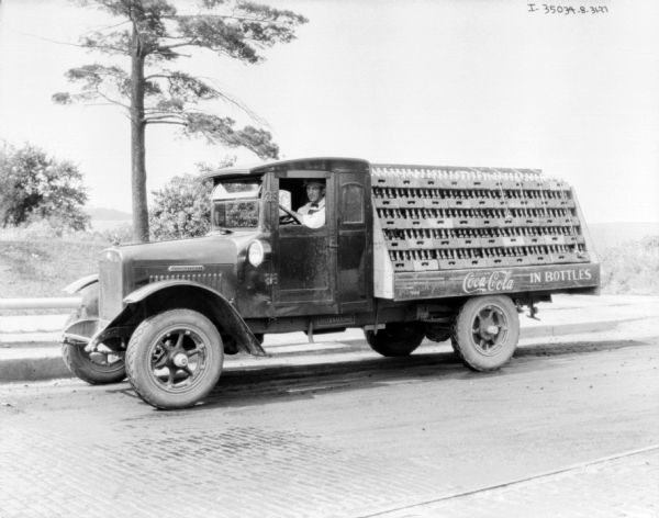 View across road towards a man sitting in the driver's seat of a Coca-Cola delivery truck parked along the opposite curb.