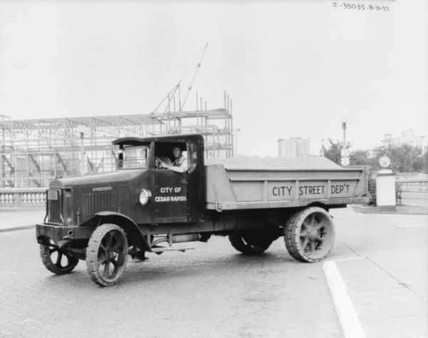 View of a man sitting in the driver's seat of a truck parked on a cobblestone street. In the background is a building under construction. The signs painted on the truck read: "City of Cedar Rapids," and "City Street Dep't."