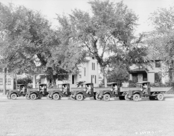 View across lawn towards men sitting in the driver's seats of a fleet of six trucks parked at an angle. A row of houses is in the background.