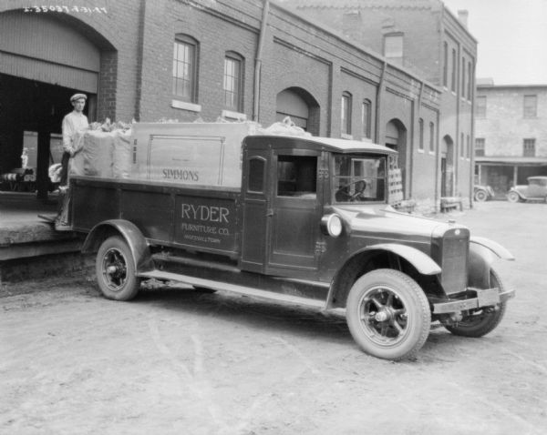 Driver's side view of a truck backed up to a loading dock. A man is standing on the loading dock in front of a brick building. The open bed of the truck is filled with bags, and a box with the sign "Simmons" printed on it. The sign painted on the truck reads: "Ryder Furniture Co. Marshalltown."