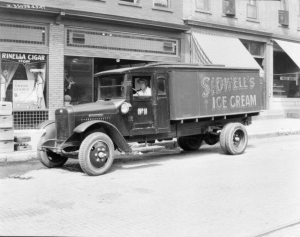 View across street towards a man sitting in the driver's seat of a truck parked along the opposite curb. There are storefronts in the background.