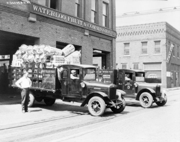 View across street towards a man sitting in the passenger side seat of a delivery truck which is backed up at the Waterloo Fruit & Commission Co. building's open garage door. The truck bed has a stake body, and is fully loaded with crates and baskets. Another man is standing on the left, and on the right a man is sitting in another delivery truck.