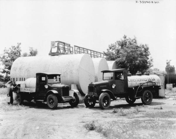 Oil delivery trucks parked near oil tanks. Signs painted on the tanks on the back of the trucks read: "Cities Service Oil Co."
