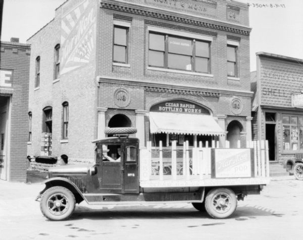 View across street towards a man sitting in the driver's seat of a delivery truck for the Hur-Mon brand of soda. The storefront in the background has a sign above the window that reads: "Cedar Rapids Bottling Works."