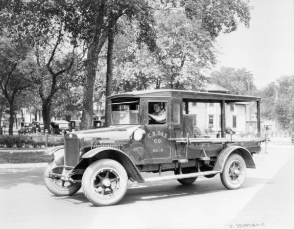 View across street towards a man sitting in the driver's seat of a C.R. Gas Co. truck. Pipes are on a rack alongside the bed of the truck.