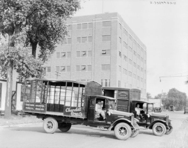 View down street towards two men sitting in two delivery trucks. A large building is in the background. A sign on the truck in front reads: "Churchill Drug Co."