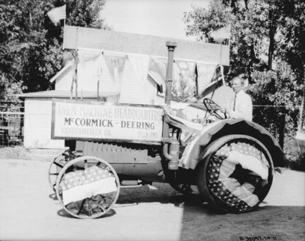 A man is driving a tractor decorated for a parade. The sign on the side reads: "Farm-Machine Headquarters, McCormick-Deering, Vandermeulen Co., Pella, Iowa."
