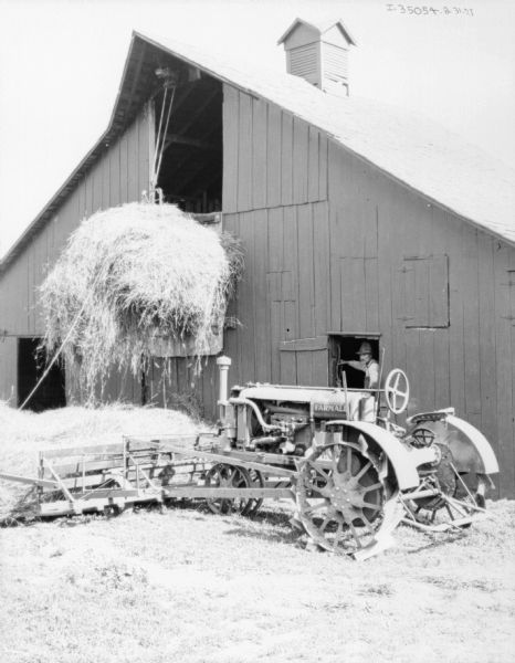 A Farmall tractor is next to a barn with a pile of hay on the ground in front of it. A man is standing in the open doorway or window of the barn, watching a load of hay attached to a pulley being moved up into the hayloft.