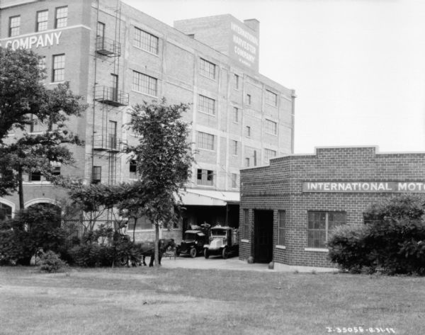 View towards the International Harvester Company plant. There is a loading dock on the right side of the building, next to a smaller brick building on the right which has a sign for Motor Trucks. Trucks are backed up to the loading dock, and a man is driving a horse-drawn wagon nearby.