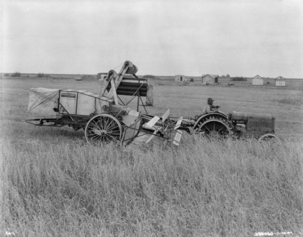 Side view of a man operating a tractor-drawn harvester-thresher in a field.