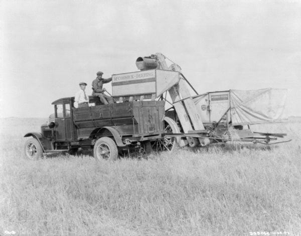 View across field towards two men sitting and standing in the back of a truck which is next to a tractor-drawn McCormick-Deering harvester-thresher.