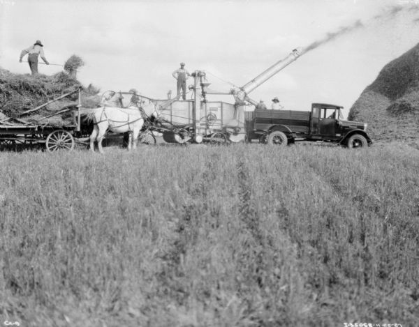 View across field towards a threshing operation. Men are standing on top of a wagon and a thresher, and the thresher is shooting hay onto a large haystack. Two men are standing in the truck bed of a truck backed up to the thresher.