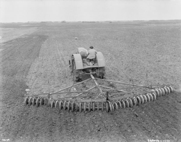 Elevated view from rear of a woman driving a tractor to pull a disk harrow in a field.