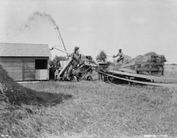 Men working on a threshing operation in the farm yard of a family farm. A haystack is in the left foreground, and on the right a man is standing on the back of a wagon feeding the threshing machine with hay. The threshing machine is being belt-driven by what is probably a tractor, which is out of frame towards the right. Two other men are standing near the thresher and a barn.