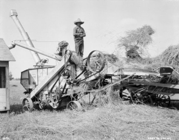 Men working on a threshing operation on a farm. A man wearing a hat is standing on top of the McCormick-Deering thresher. Another man on the right is standing on a wagon feeding the threshing machine with hay. There is a barn on the left. The threshing machine is being belt-driven by what is probably a tractor, which is out of frame towards the right.
