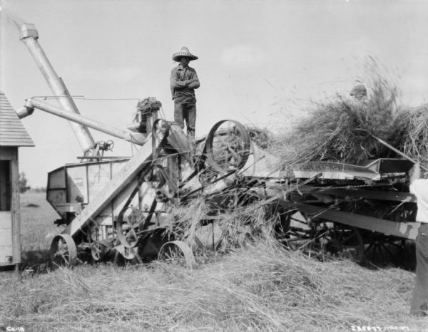 Men working on a threshing operation on a farm. A man wearing a hat is standing on top of the McCormick-Deering thresher. Another man on the right is standing on a wagon feeding the threshing machine with hay. There is a barn on the left. The threshing machine is being belt-driven by what is probably a tractor, which is out of frame towards the right.