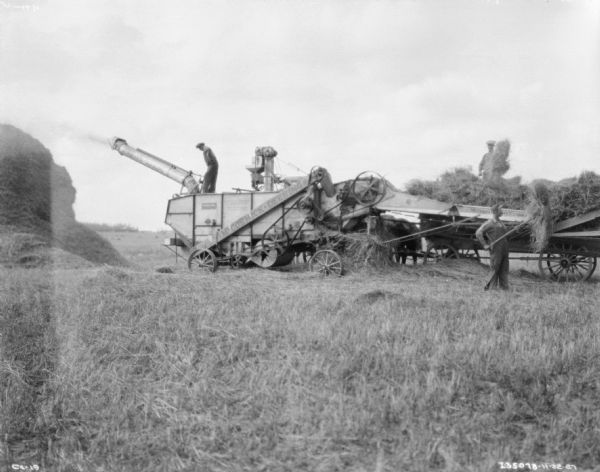 View across field towards men working on a threshing operation on a family farm. A man is standing on top of a McCormick-Deering threshing machine, and two men on the right are standing near a wagon loaded with hay.