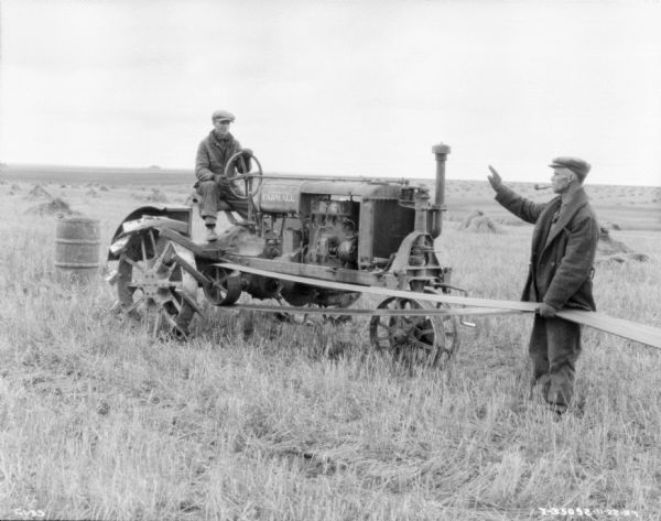 A man is sitting on a Farmall tractor, and another man is standing on the right in front of the tractor holding the belt attached to the tractor. The machinery being belt-driven by the tractor is out of frame. In the background are stacks of harvested grain in the field.