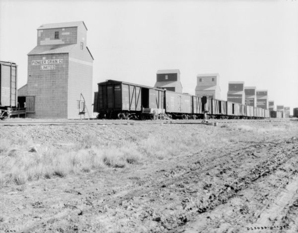 A long line of grain elevators are behind railroad tracks. Railroad cars are sitting on the tracks.