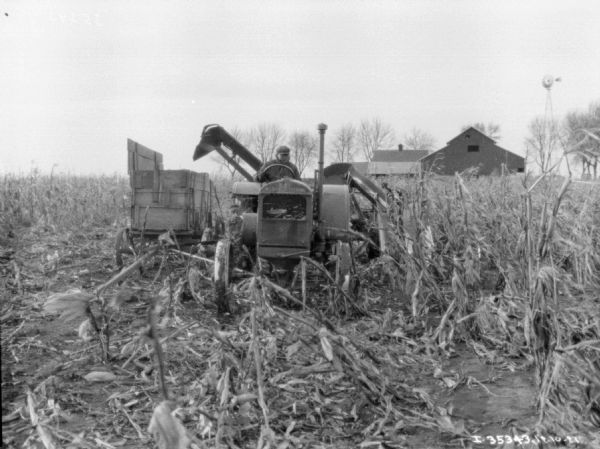 View from front of a man on a Farmall tractor with a corn picker working in a field. There is a wagon on the left. Farm buildings are in the background.