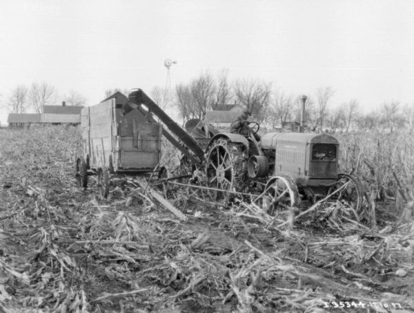 View from front of a man on a Farmall tractor with a corn picker working in a field. There is a wagon on the left. Farm buildings are in the background.