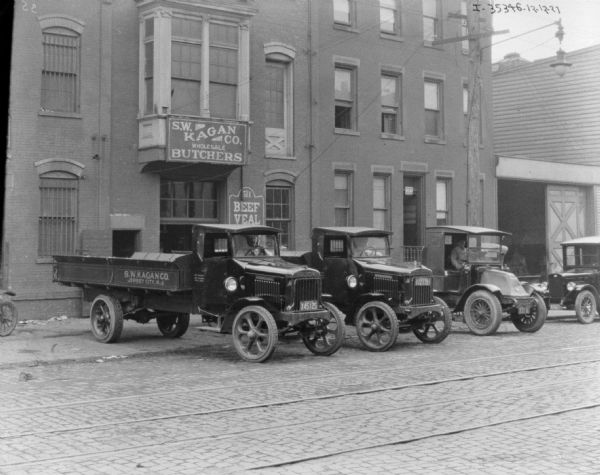 View across street towards a fleet of three meat delivery trucks backed up to a building with a sign for: "S.W. Kagan Co. Wholesale Butchers." Three men are sitting in the driver's seat of each truck.