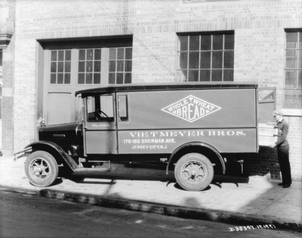 View from street towards a man loading bread into the back of a delivery truck. Three small American flags are attached to the front of the hood.