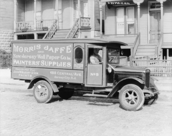 View across cobblestone street towards a man sitting in the driver's seat of a delivery truck. The sign painted on the truck reads: "Morris Jaffe and New Jersey Wall Paper Co. Inc. Painters' Supplies."