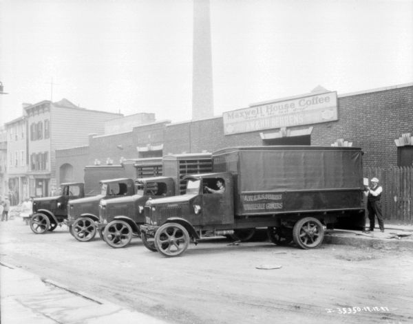 Fleet of grocery delivery trucks backed up to a building with a sign that reads: "A.W. & H.H. Behrens Wholesale Grocers." One man is sitting in the driver's seat of the nearest truck, and another man is standing at the back of the truck.
