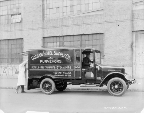 View across street towards a man sitting in the driver's seat of a delivery truck. The sign painted on the truck reads: "Gotham Hotel Supply Co., Inc. Purveyors Hotels Restaurants Steamships."