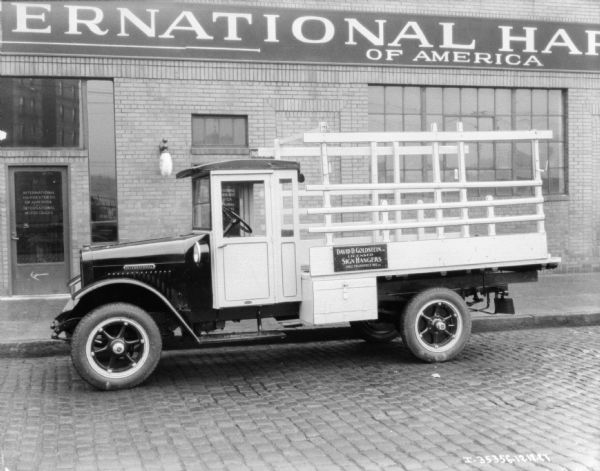 View across street towards a truck parked on a cobblestone street along the curb. The sign on the truck reads: "David D. Goldstein Licensed Sign Hangers." The building in the background has a sign for International Harvester of America.