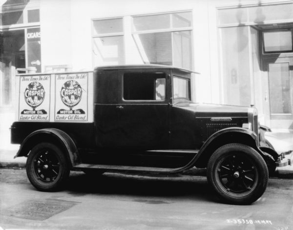 View across street towards a delivery truck parked along the curb. The sign on the truck reads: "Three Times the Life! Caspar Motor Oil." There is a store in the background with a sign on the window that reads: "Cigars."
