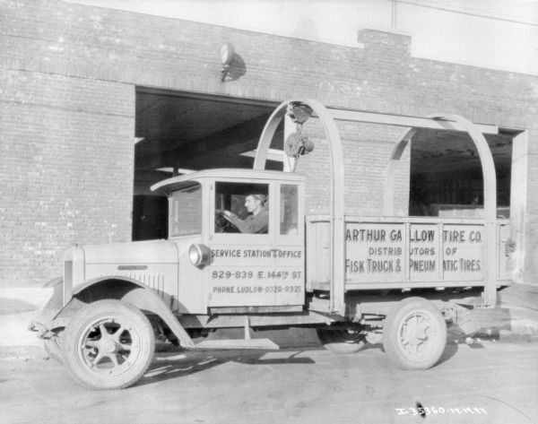 View towards a man sitting in the driver's seat of a delivery truck. In the background is a brick building with large, open garage doors. The sign on the truck reads: "Arthur Gallow  Tire Co. Distributors of Fisk Truck & Pneumatic Tires."