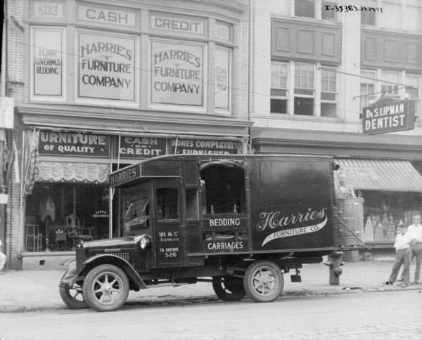 View across street towards a delivery truck parked along the curb. The sign on the truck reads: "Harries Furniture Co." The truck is parked in front of a Harries Furniture Co. storefront, and a man is standing in the back of the truck with a piece of furniture.