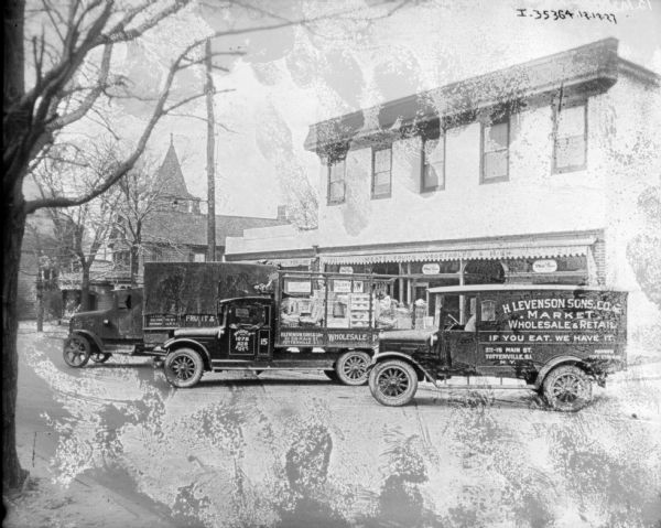 View across street towards a fleet of trucks parked at an angle in front of storefronts. The sign on the trucks reads: "H. Levenson Sons & Co. Inc. • Market • Wholesale & Retail, If You Eat It We Have It."