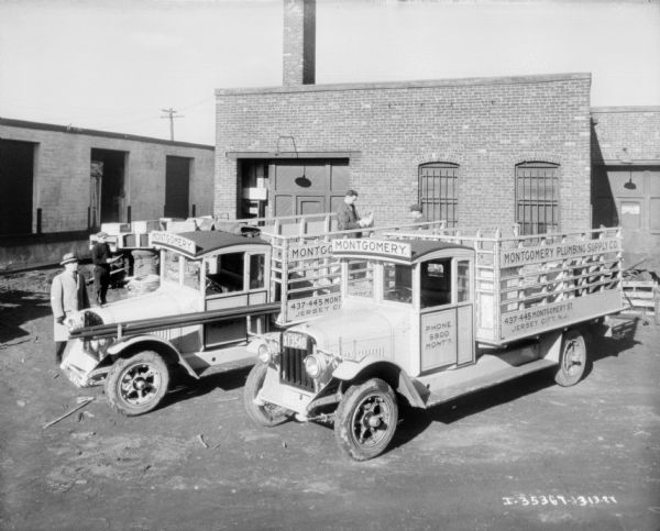Elevated view of delivery trucks parked in front of a brick building. Men are standing near the trucks, and also standing in the truck beds, which have stake bodies. The signs on the trucks read: "Montgomery Plumbing Supply Co."